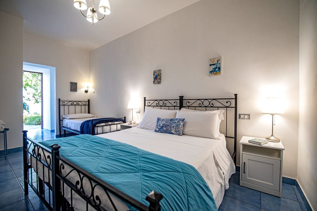 A bright, spacious bedroom with two wrought-iron beds dressed in white linens and blue accents, decorated in Mediterranean style with pale walls and access to an outdoor terrace.