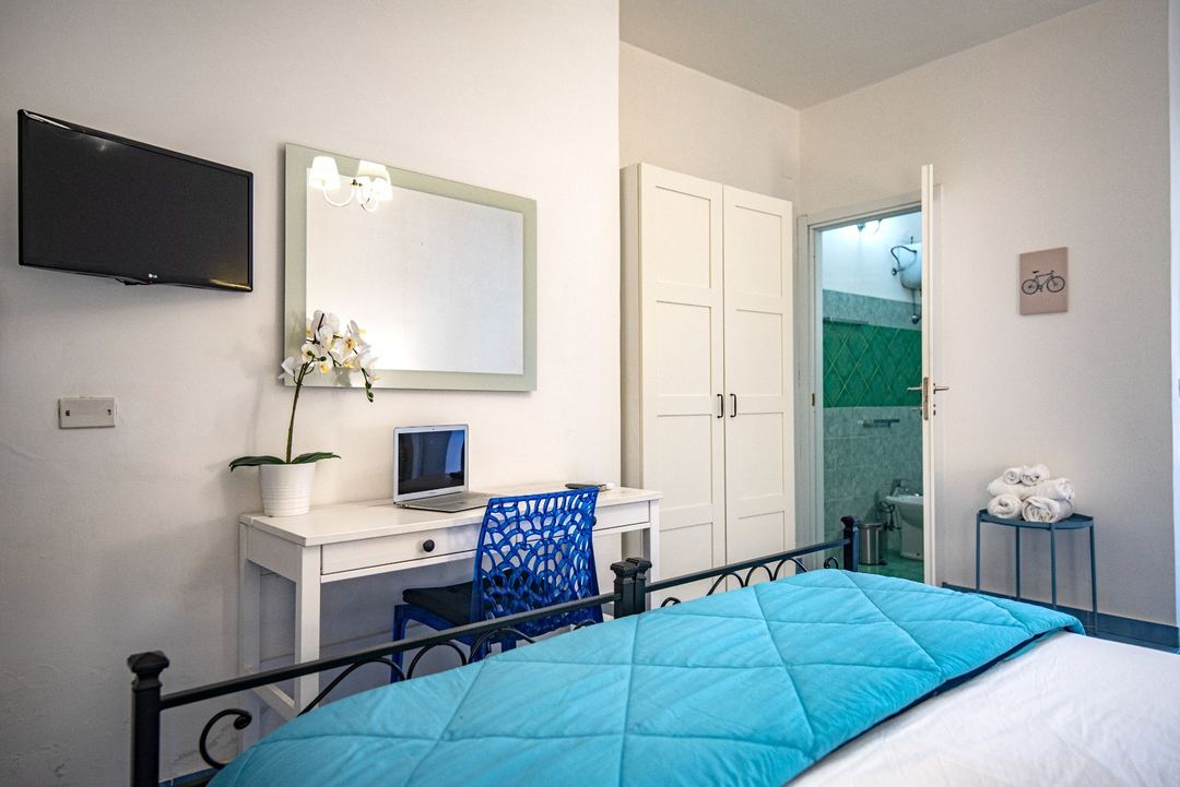 A bright guest bedroom with a blue quilted bed, white desk with blue chair, mirrored wall, wall-mounted TV, and an ensuite bathroom visible through an open door.