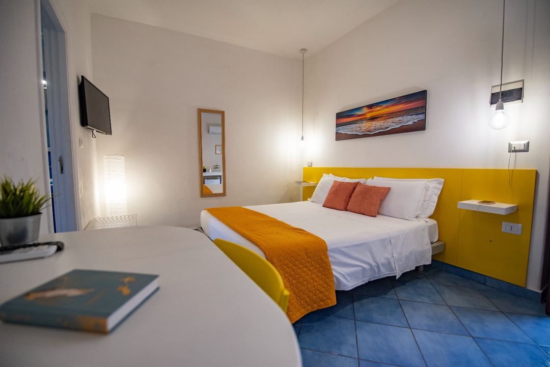 A bright modern bedroom with a yellow headboard, white bedding, orange throw, and coastal artwork, featuring blue tile flooring and contemporary pendant lighting.