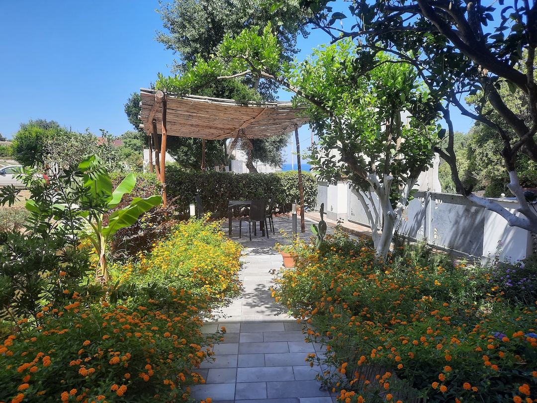 A Mediterranean garden pathway with vibrant orange marigolds leading to a wooden pergola-covered terrace overlooking the sea.