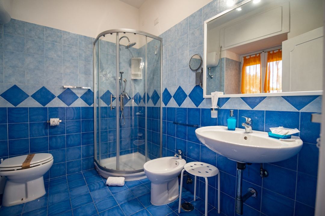 A bright bathroom with blue tile walls, curved glass shower enclosure, white fixtures, and a pedestal sink with large mirror.