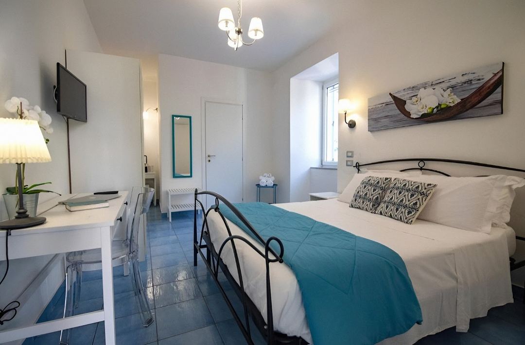 A bright, modern hotel bedroom with a teal accent color, wrought-iron bed frame, work desk, and nautical decor.