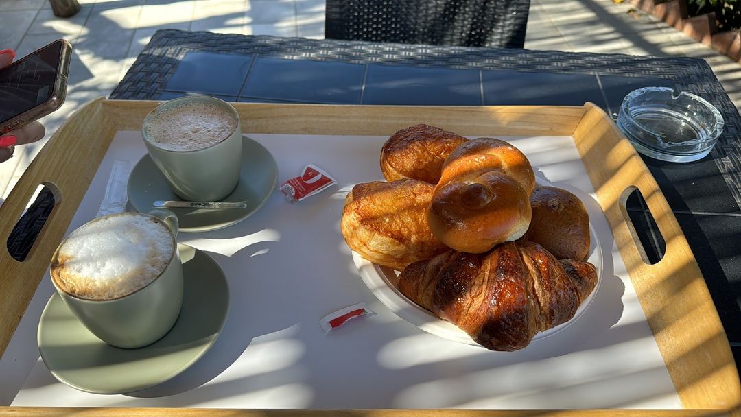 A breakfast tray featuring two cappuccinos, fresh croissants, and pastries served on a wooden table in bright sunlight.