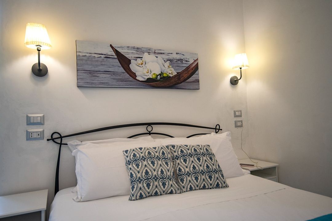 A neatly made bed with white pillows and a patterned cushion, framed by a black iron headboard, with wall-mounted sconces and decorative artwork above.